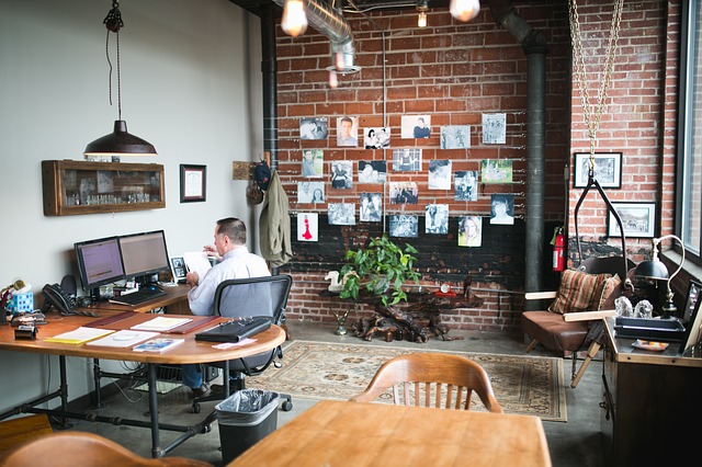 Kanceláře se zděnou zdí, mnoho obrázků, stůl, počítače a muž co pracuje